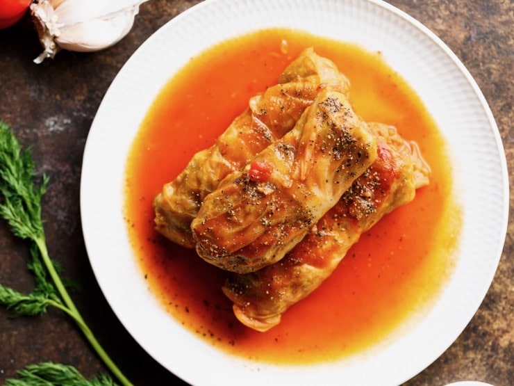 Three stuffed cabbage leaves - holishkes on a plate with sauce, fresh dill and garlic on the side.