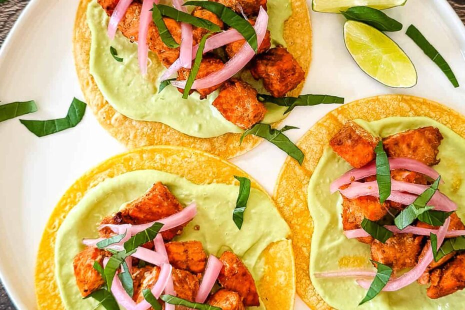 Air fryer salmon tacos on corn tortillas topped with avocado lime crema, pickled red onions, and fresh cilantro, served with lime wedges on a white plate.