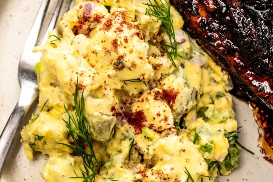 A plate with creamy potato salad garnished with dill and paprika sits beside a piece of grilled, glazed meat and a fork.