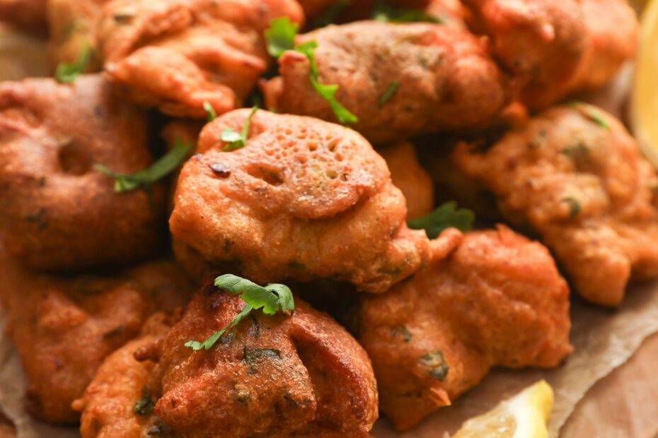 Close-up of crispy, golden-brown pakora (fritters) garnished with fresh coriander, served on a rustic wooden board with a wedge of lemon and chutney in the background.