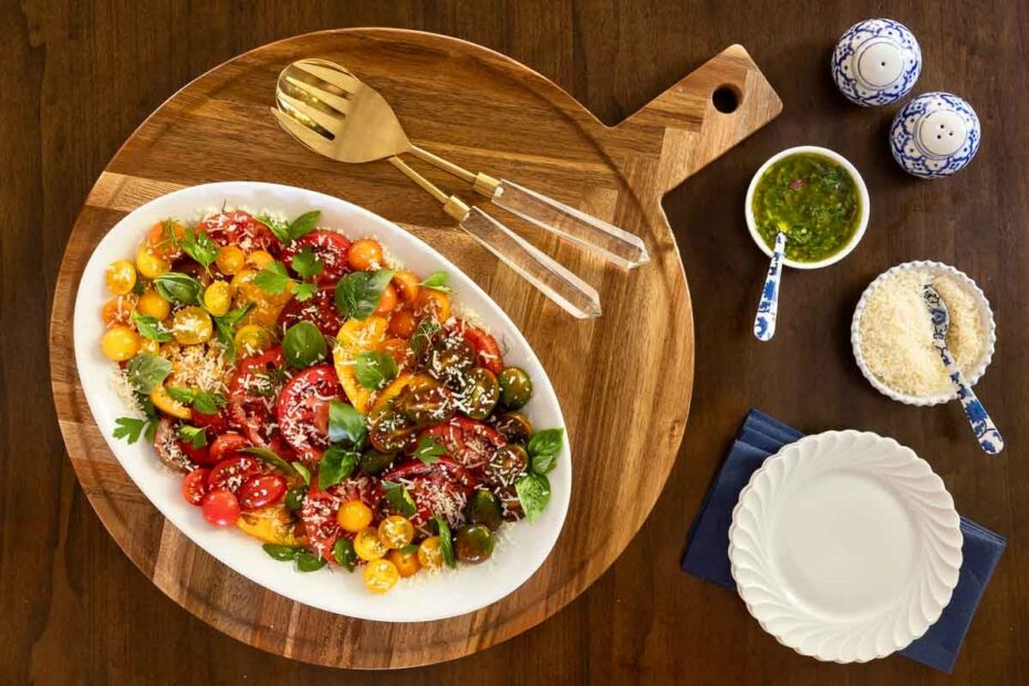 Heirloom cherry tomato salad with fresh herbs and grated Parmesan on a white platter, served on a wooden board with vinaigrette and extra cheese on the side.