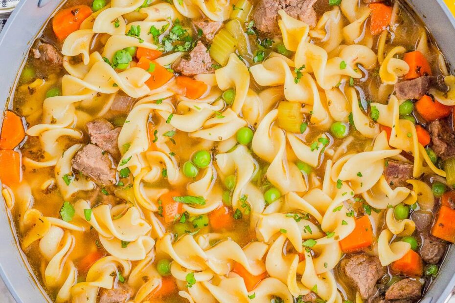 A pot of beef noodle soup with egg noodles, carrots, peas, celery, and chunks of beef, garnished with parsley. Uncooked pasta and herbs are scattered on the white surface nearby.