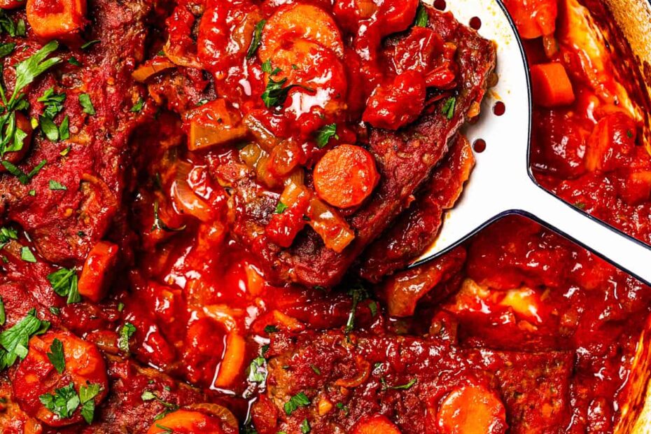A spoon lifting Swiss steak from tomato gravy in a Dutch oven.