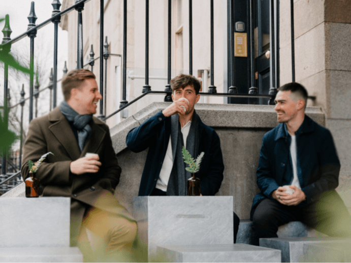 Tadhg Guiry sits outside in a coat and scarf drinking from a paper cup, in between his CSM colleagues.