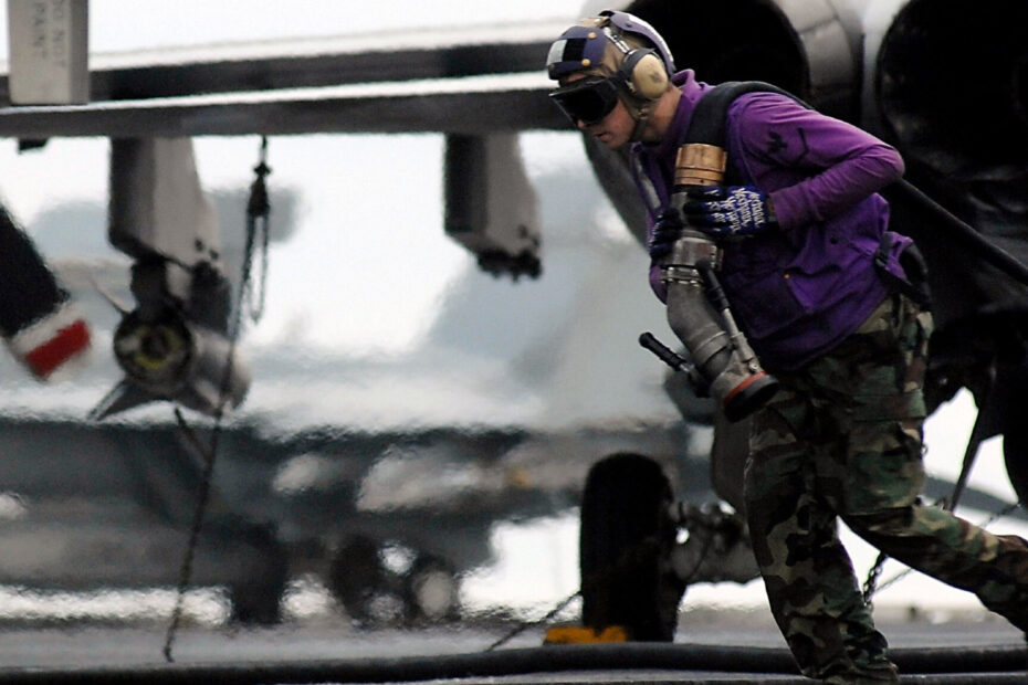 A man wearing a pilot's helmet and sunglasses and a purple jacket and camouflage pants carries a large hose away from a plane.