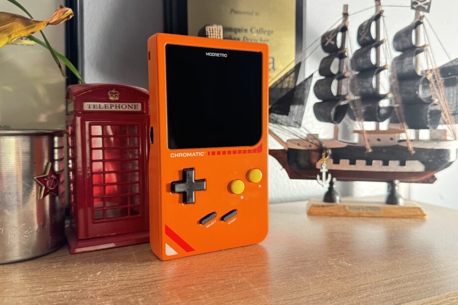 An orange ModRetro Chromatic on a shelf next to a model pirate ship and a model red English phone booth.