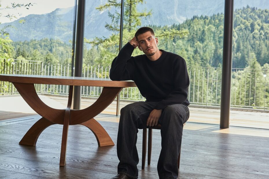 A man dressed in black sits at vintage style table with large windows behind and forests outside.