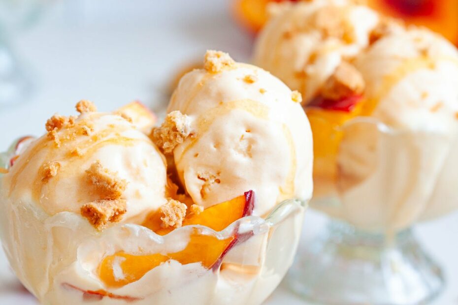 two glass sundae bowls with scoops of peach ice cream with peach slices and cookie crumbles