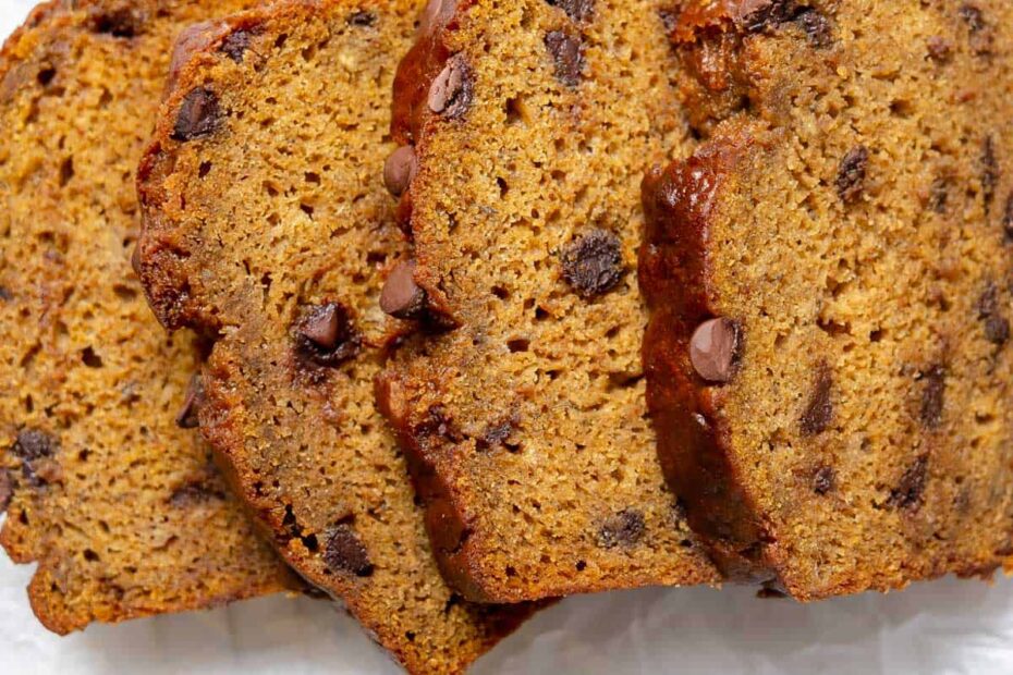 Three slices of chocolate chip pumpkin banana bread are arranged on parchment paper, with a few loose chocolate chips and part of a banana visible nearby.