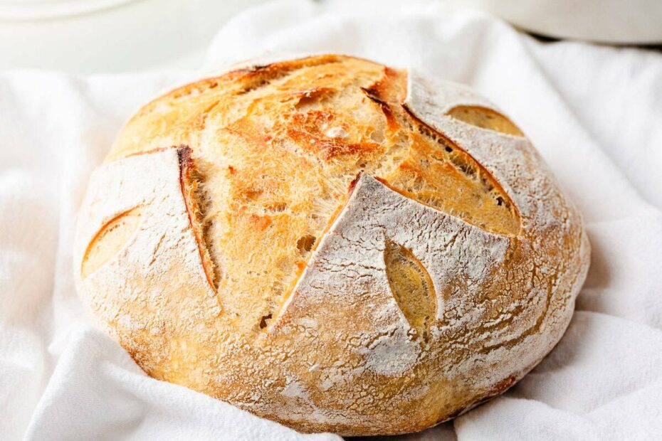 Golden brown round loaf of same day sourdough bread with decorative scoring, placed on a soft white kitchen towel.