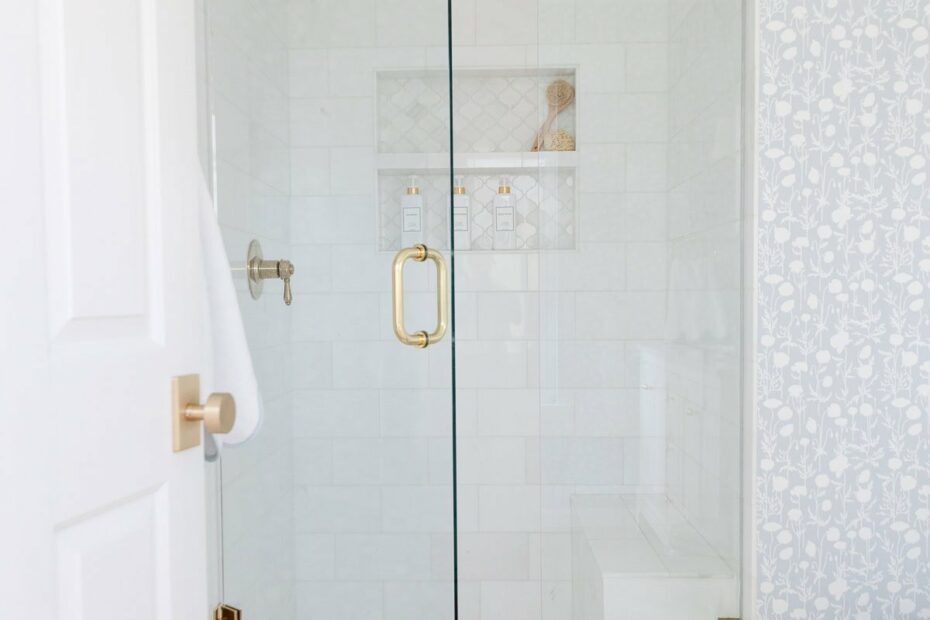 Bright bathroom with a glass shower enclosure, gold fixtures, white tile, a patterned wall, and a scalloped white bath mat on a light wood floor.
