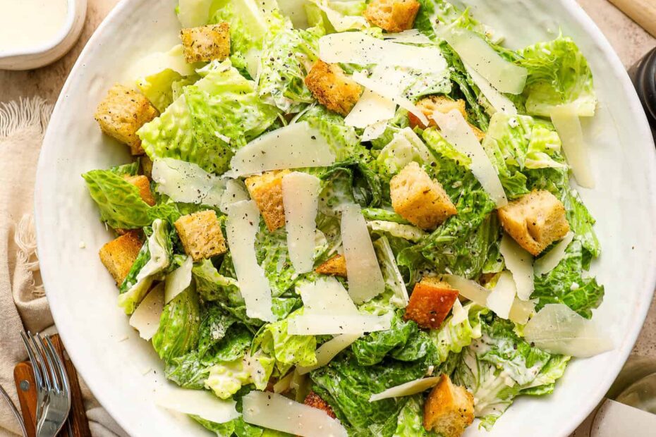 A bowl of classic Caesar Salad with romaine lettuce, croutons, and shaved parmesan cheese sits on a beige table set with utensils, wine glasses, and cheese.