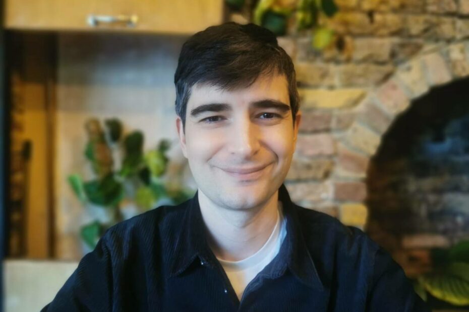 A selfie of Louis Barson in front of a stone fireplace wearing a black shirt and smiling at the camera.