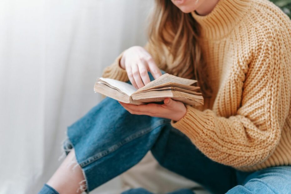 A woman in a cozy mustard sweater reads an old book by the window, illustrating what one small change looks like in real life through quiet reflection and growth.