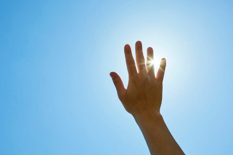 A hand reaching toward the bright sun in a clear blue sky, with sunlight shining between the fingers, symbolizing clarity, hope, and guidance.