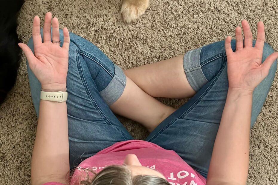 woman sitting with legs crossed on the floor with her palms open, face up, resting on her knees. There is a dog sitting next to her, partially in the picture.