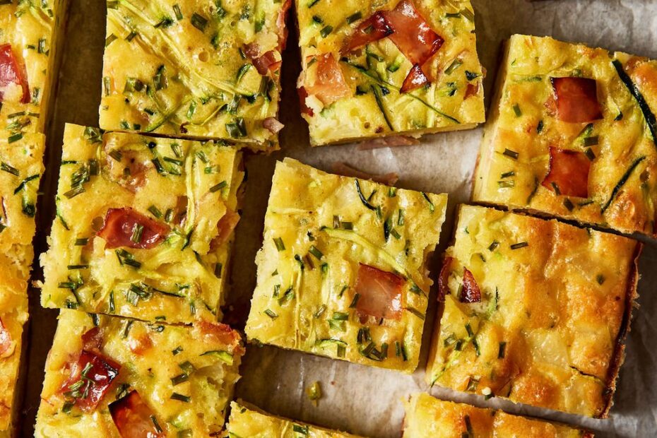 Sourdough zucchini slice made with sourdough discard, grated zucchini, cheddar cheese, ham, chives and eggs, baked and cut into squares on a parchment-lined, wooden cutting board.