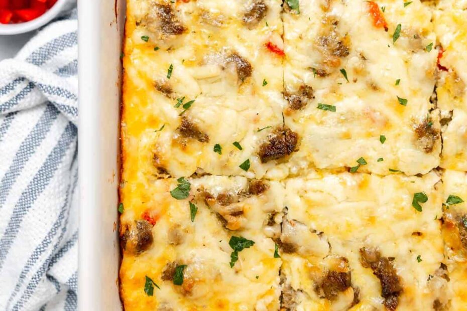 A baked breakfast casserole with sausage, cheese, and vegetables cut into squares, in a white dish on a striped towel, with a small bowl of diced red bell peppers nearby.