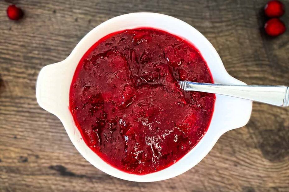 A white bowl filled with homemade cranberry sauce on a wooden table, surrounded by fresh cranberries.