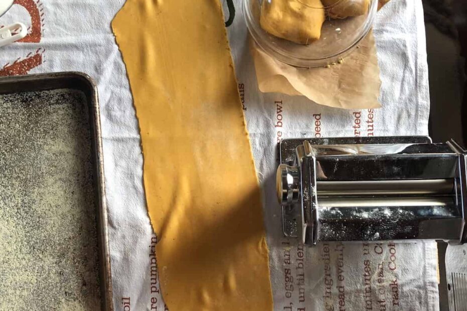 Homemade pumpkin pasta dough with lasagna sheets and pasta machine next to it with 3 pumpkin pasta dough balls covered in the background.