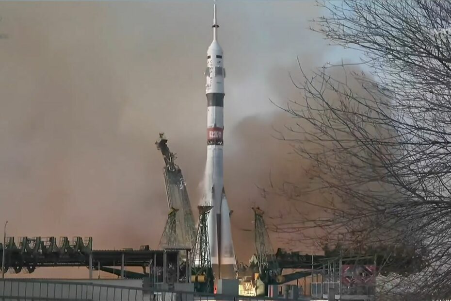 A white rocket is pictured lifting-off from a launch site in Russia. Fiery exhaust and smoke is visible pouring from the rocket's engines as it lifts into a grey sky.