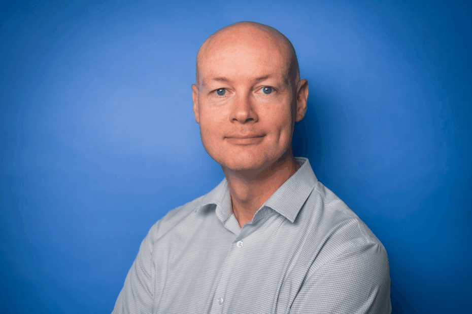 Caucasian male with an open necked shirt on a blue background.
