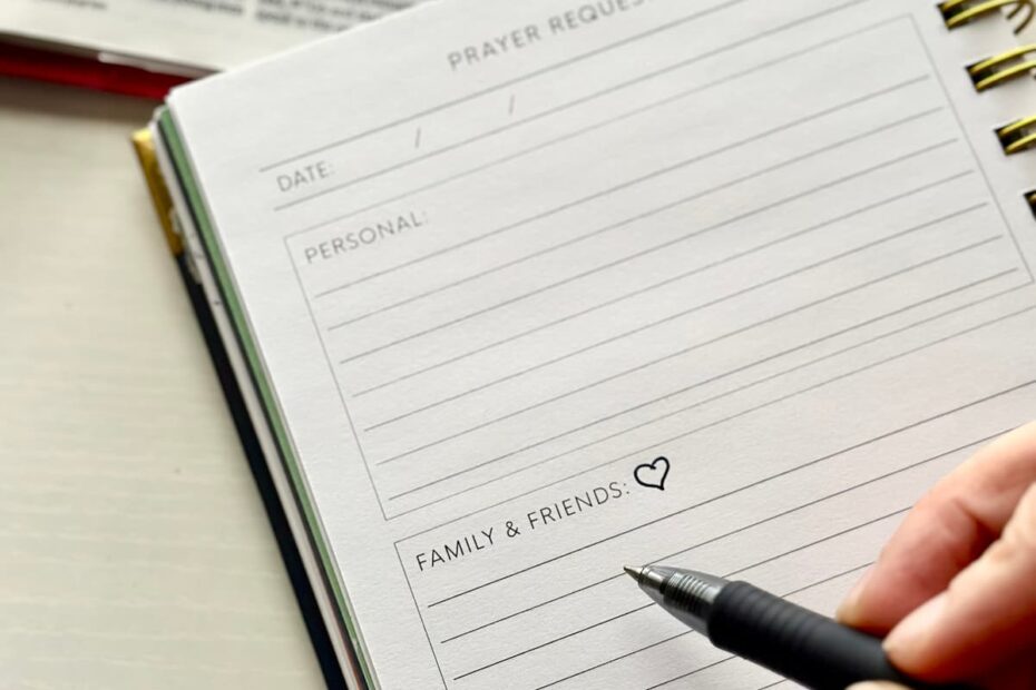 Hand holding a pen above a prayer journal with a heart drawn next to "friends & family" on the page. A Bible is open on the desk next to the notebook