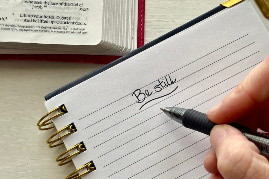 Prayer journal with "Be still" written on the page with a hand holding a pen and Bible open next to the spiral journal