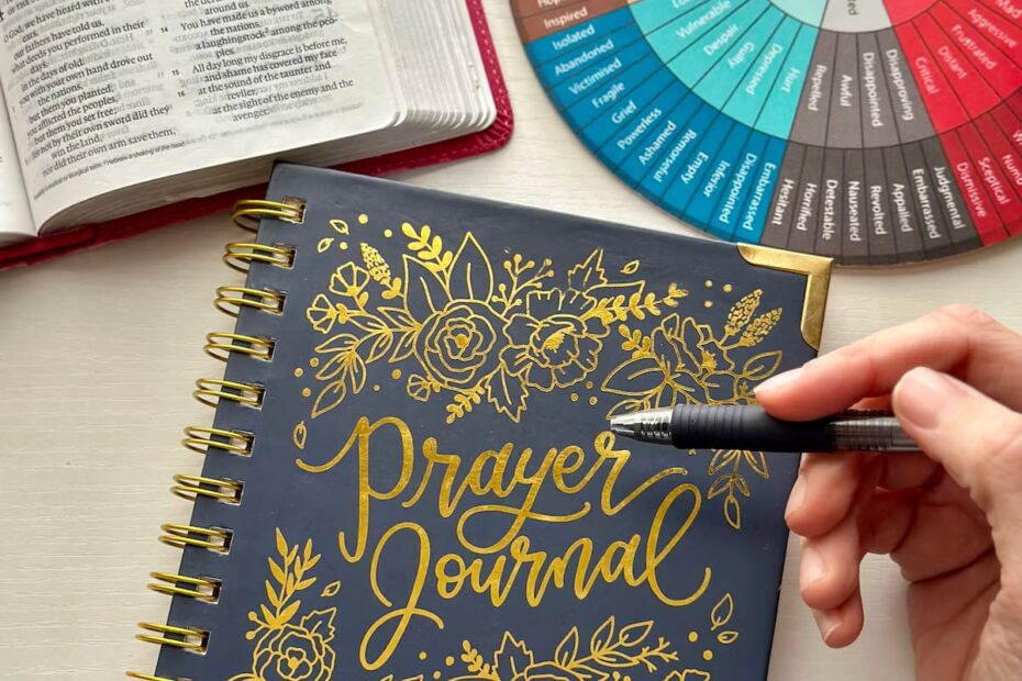 A desk with an open Bible, a feelings/emotions wheel, a prayer journal, and a hand holding a pen