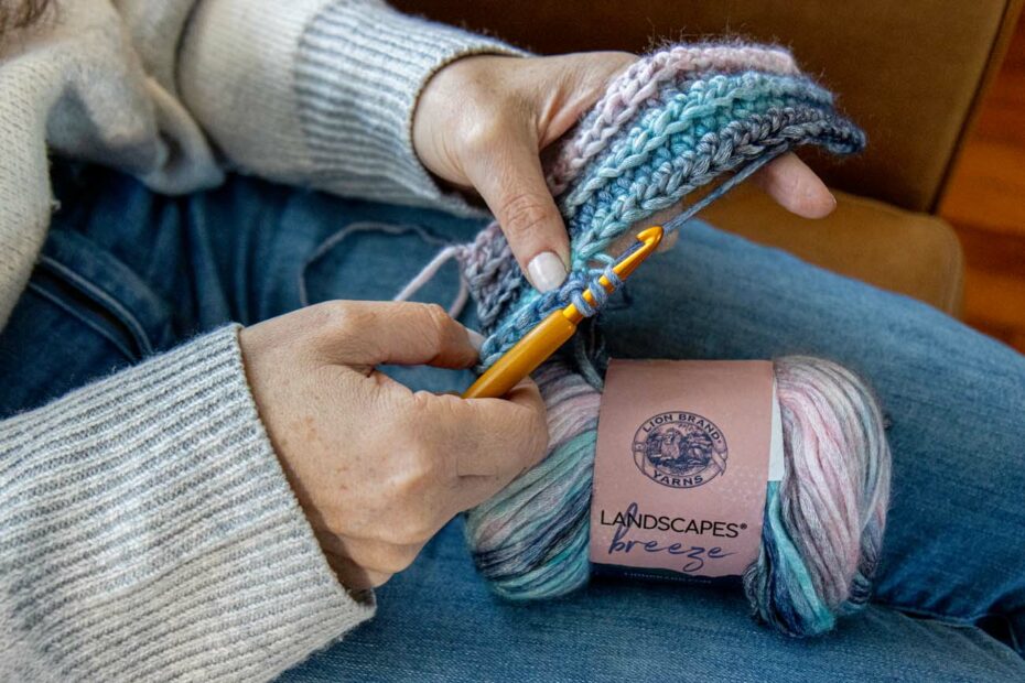 Hands crocheting with gold hook and Lion Brand Landscapes Breeze yarn in coastal tones, showing active stitching technique with yarn skein resting on denim-clad lap