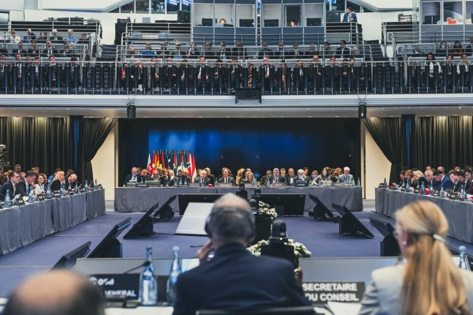 A series of people sit at four different tables making a square to face each other with various flags of their corresponding countries next to them.