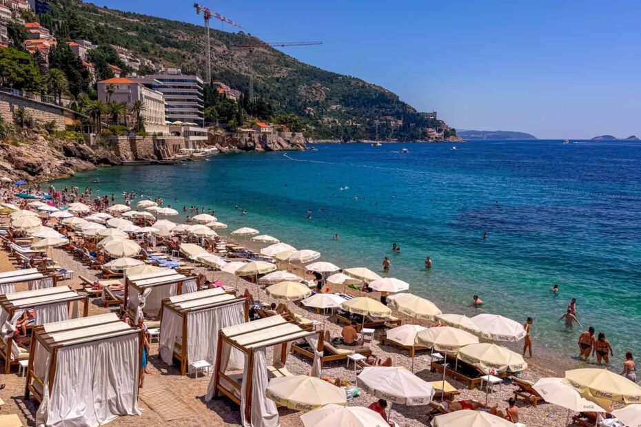 Crowded beach with rows of lounge chairs and umbrellas, people sunbathing and swimming in the clear blue sea—this spot often features in the Dubrovnik beach guide and is among the best beaches near Old Town Dubrovnik, with scenic hills and buildings in the background.