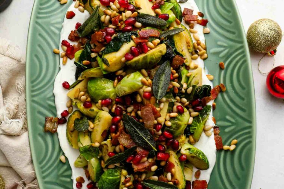 Holiday Brussels Sprouts plated with bacon, pine nuts, pomegranate seeds, and fried sage on a bed of yogurt, served on an oval green dish.