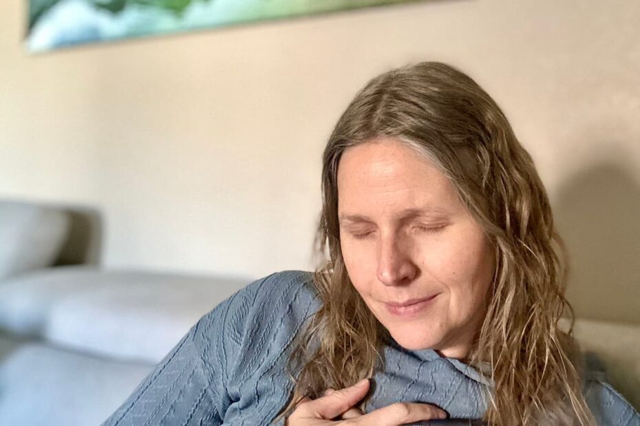 Woman sitting on her couch with her eyes closed hands gently folded over her heart, and eyes closed with a slight smile on her face as she prays