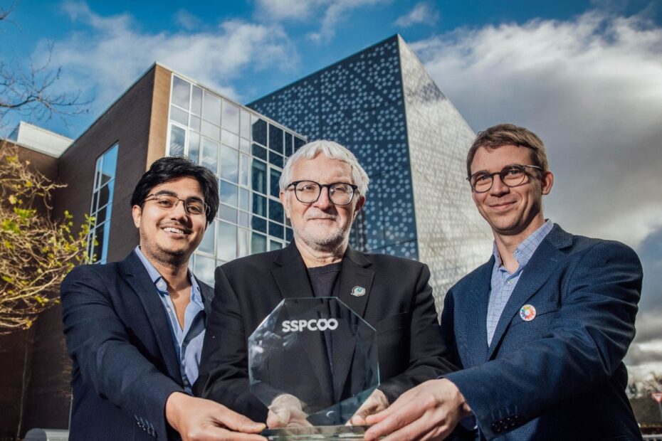 Three men stand close together holding a glass award in front a large glass building on the UL campus.