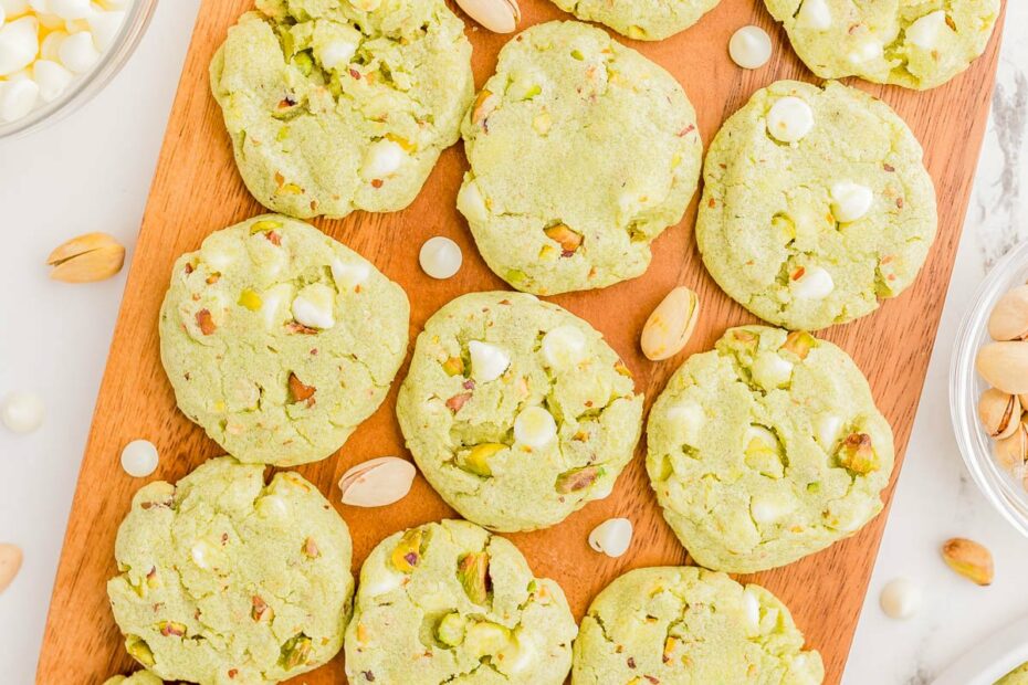 A wooden board with 16 green pistachio cookies topped with white chocolate chips, surrounded by scattered pistachios and a bowl of white chocolate chips.