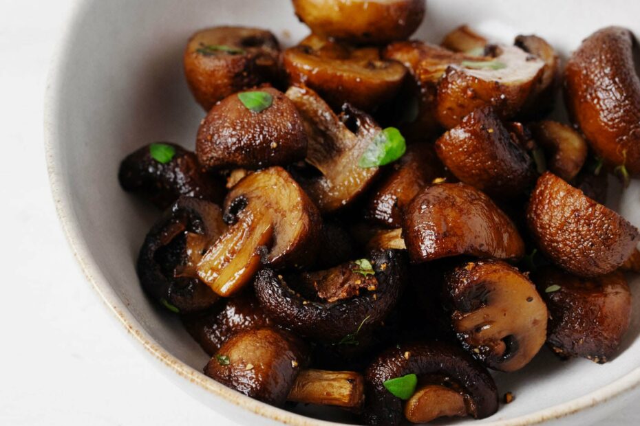 An asymmetrical white bowl is holding deep brown, air fryer mushrooms. The mushrooms are dotted with bright green herbs.