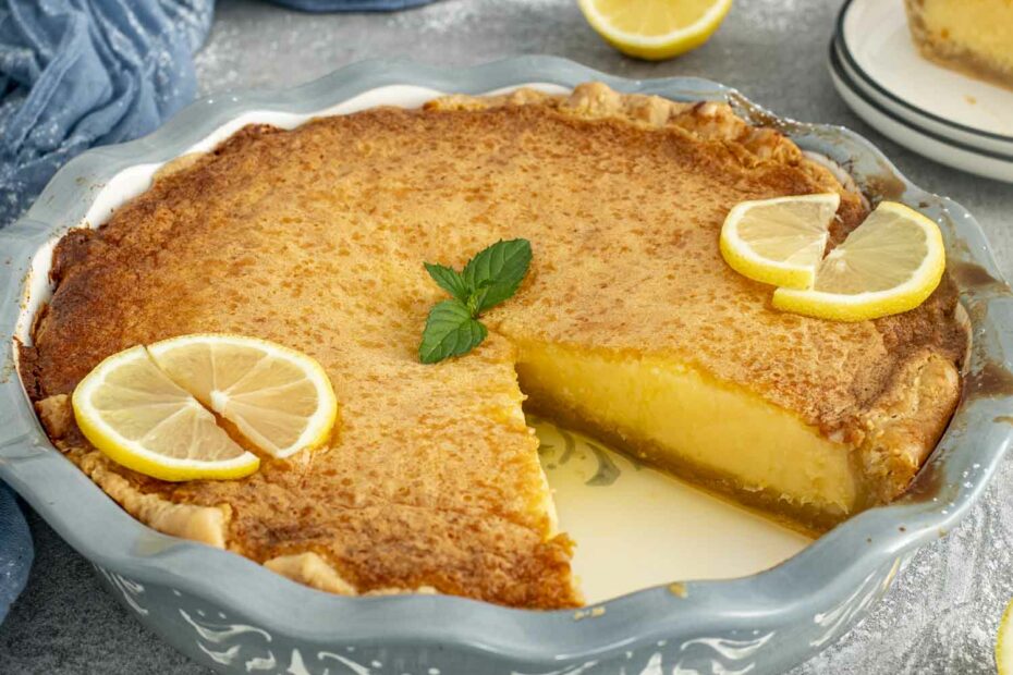 Whole lemon chess pie with slice removed, topped with lemon slices and mint, showing its smooth custard and golden crust.