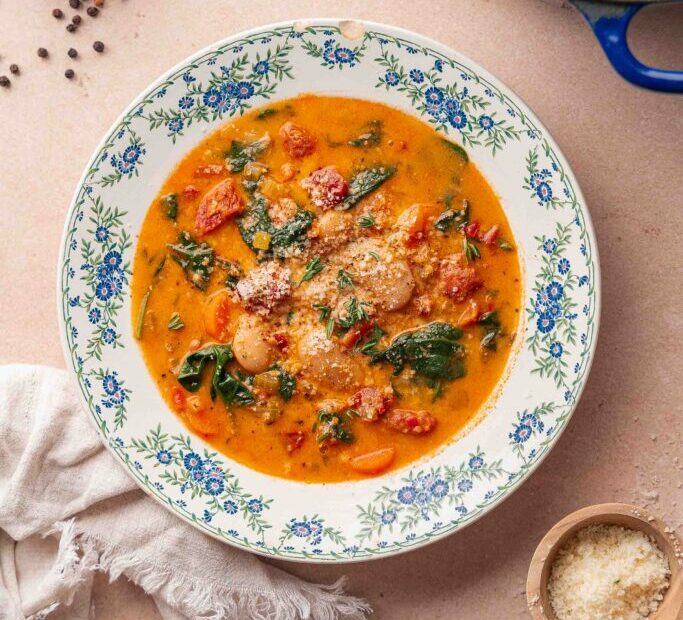 A bowl of creamy chorizo soup with spinach, beans and carrots is served on a flower-bedecked plate, sprinkled with grated cheese and accompanied by a cloth napkin.