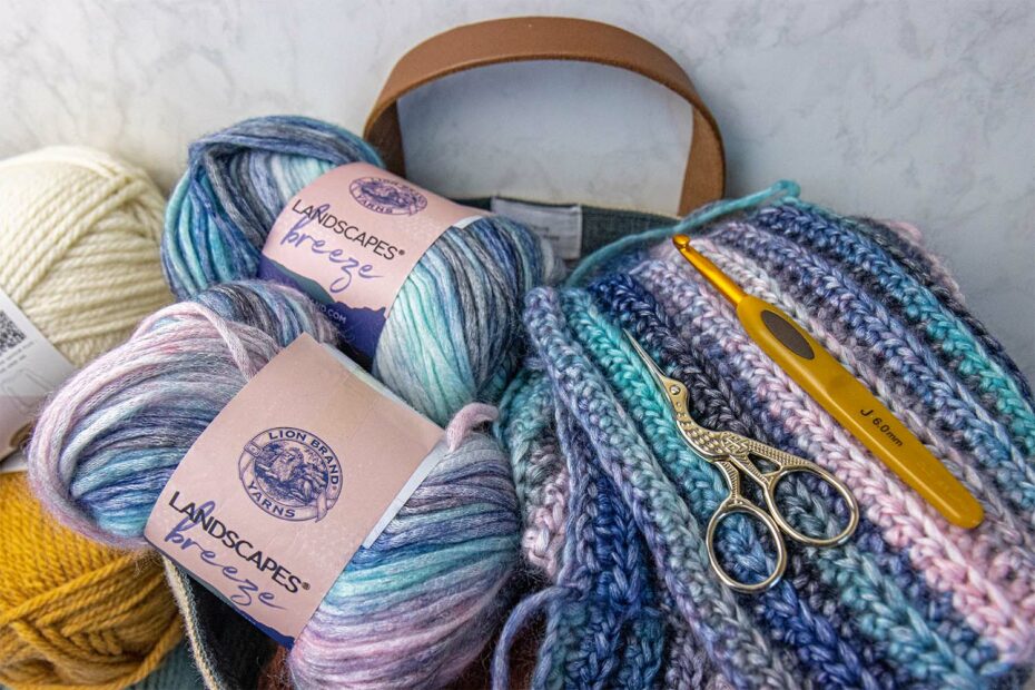 Lion Brand Landscapes Breeze gradient yarn skeins in coastal blues, pinks, and creams with completed crochet squares, gold scissors, and yellow crochet hook in canvas tote bag for our Lion Brand Yarn Review