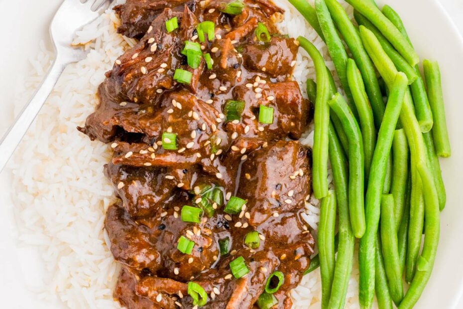 A plate of white rice topped with shredded beef in a dark sauce, garnished with green onions and sesame seeds, served with green beans and a fork on the side.
